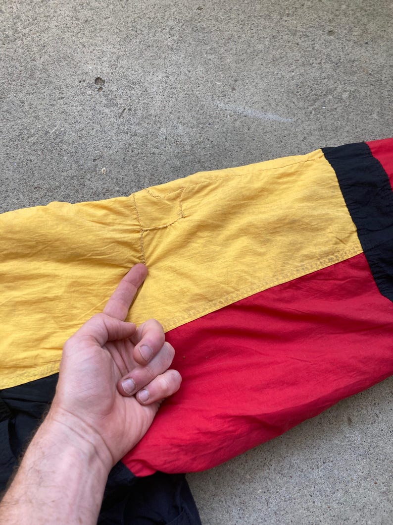 May include: Close-up of a section of a colorful jacket. The jacket features a color block design with yellow, red, and black panels. The fabric appears to be a lightweight, possibly nylon material. The stitching and seams are visible.