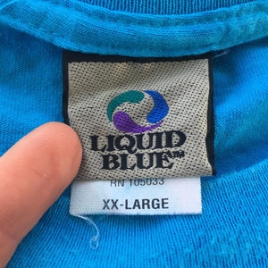Może przedstawiać: Niebieska koszulka z białą metką, na kt&oacute;rej widnieje napis "Liquid Blue" i "RN 105033" wraz z wirującym niebiesko-fioletowym logo. Metka zawiera r&oacute;wnież napis "XX-Large".