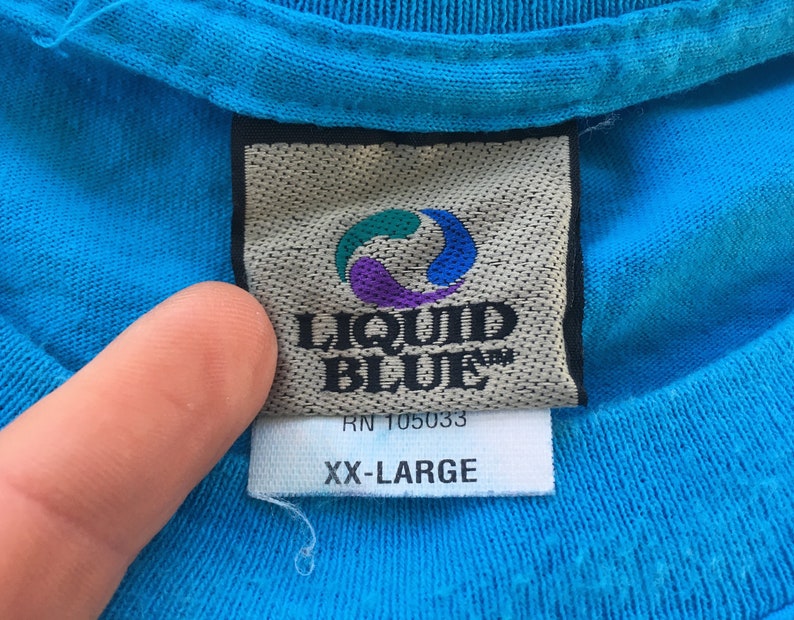 Może przedstawiać: Niebieska koszulka z białą metką, na kt&oacute;rej widnieje napis "Liquid Blue" i "RN 105033" wraz z wirującym niebiesko-fioletowym logo. Metka zawiera r&oacute;wnież napis "XX-Large".