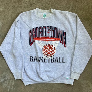 Puede incluir: Una sudadera gris claro de cuello redondo con un gráfico de una canasta de baloncesto con un balón dentro. El texto GEORGETOWN está arqueado sobre la canasta en azul marino y rojo, con la palabra BASKETBALL impresa en letras negras debajo.
