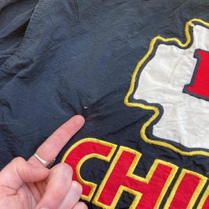 May include: Black jacket with the word "CHIEFS" embroidered in red and yellow. The jacket also features a white and red logo. The jacket is made of a shiny material.