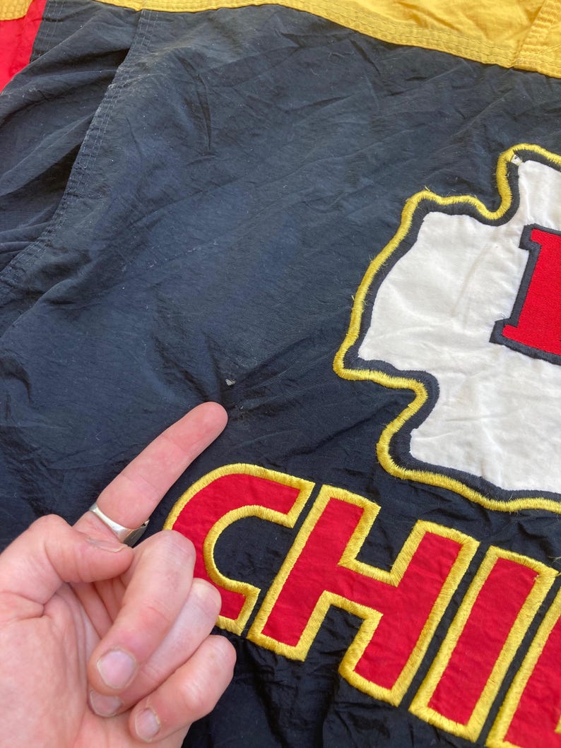 May include: Black jacket with the word "CHIEFS" embroidered in red and yellow. The jacket also features a white and red logo. The jacket is made of a shiny material.