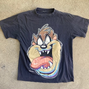 May include: Black t-shirt featuring a cartoon image of Taz the Tasmanian Devil. The image is in a colourful style with a light blue background.