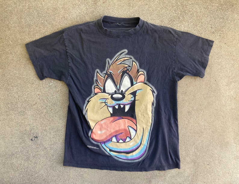 May include: Black t-shirt featuring a cartoon image of Taz the Tasmanian Devil. The image is in a colourful style with a light blue background.