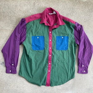 May include: A long-sleeved button-down shirt with a color-blocked design. The shirt is purple on the sleeves, red on the top, green in the middle, and blue on the pockets.