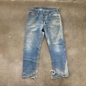 Puede incluir: Vaqueros de mezclilla azul descolorido con detalles desgastados, incluyendo rasgaduras y marcas de uso. Los vaqueros tienen bragueta con botones y diseño clásico de cinco bolsillos. La mezclilla muestra signos de edad y uso.