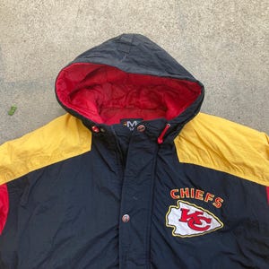 May include: A hooded jacket with a black body, yellow shoulders, and red accents. The jacket features the Kansas City Chiefs logo and the word "CHIEFS" in red and white.