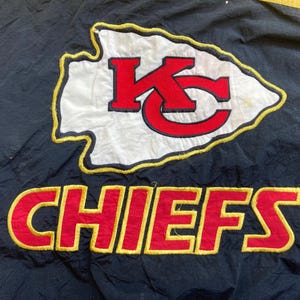 May include: Black fabric with a large embroidered logo of the Kansas City Chiefs. The logo features a white arrowhead with a red "KC" and the word "CHIEFS" in red with a yellow outline.