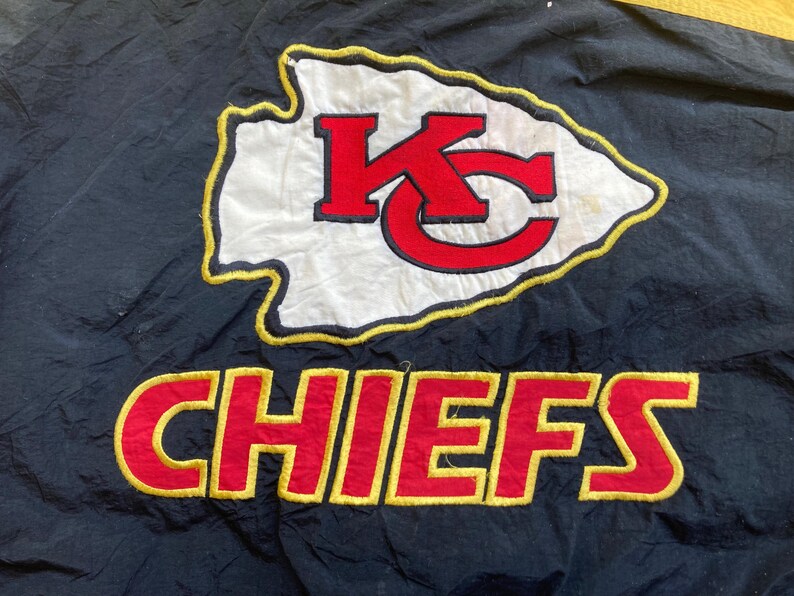 May include: Black fabric with a large embroidered logo of the Kansas City Chiefs. The logo features a white arrowhead with a red "KC" and the word "CHIEFS" in red with a yellow outline.