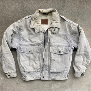 Puede incluir: Una chaqueta vaquera gris con forro de sherpa y dos bolsillos grandes en la parte delantera. La chaqueta tiene cierre de cremallera y cuello abotonado.