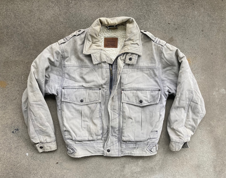 Puede incluir: Una chaqueta vaquera gris con forro de sherpa y dos bolsillos grandes en la parte delantera. La chaqueta tiene cierre de cremallera y cuello abotonado.