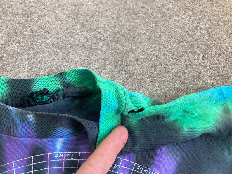 Puede incluir: Primer plano del cuello de una camiseta tie-dye con un esquema de color negro y verde. La camiseta tiene un peque&ntilde;o desgarro cerca del escote.