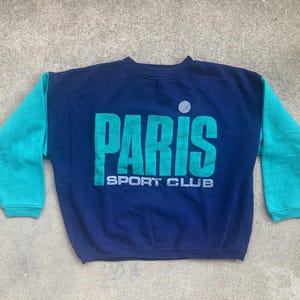 Può includere: Una felpa blu navy vintage con maniche turchese. La parte anteriore presenta la scritta "PARIS" in grandi lettere turchesi sopra "SPORT CLUB". Un piccolo cerchio bianco è sopra la lettera "I".