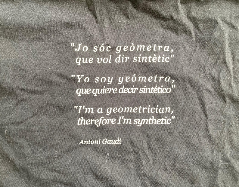 Puede incluir: Camiseta negra con texto blanco en tres idiomas: catal&aacute;n, espa&ntilde;ol e ingl&eacute;s. La cita, atribuida a Antoni Gaud&iacute;, dice: &laquo;Soy ge&oacute;metra, por lo tanto soy sint&eacute;tico&raquo;.