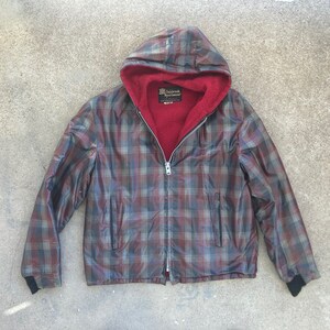 May include: A plaid hooded jacket with a red fleece lining. The jacket has a zip closure and black trim on the cuffs and waistband. The label on the jacket reads "The Sportsman Apparel Co."