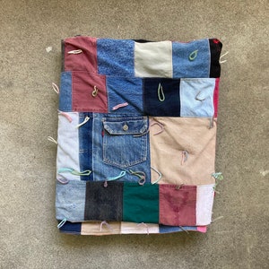 May include: A patchwork cushion made from various denim and fabric scraps. The design includes a denim jacket pocket and is stitched together with colorful thread. The cushion is square and rests on a concrete surface.