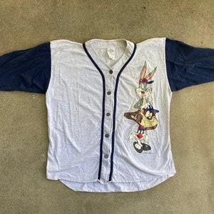 Puede incluir: Una camiseta de béisbol gris y azul con una imagen de dibujos animados de Bugs Bunny y el Demonio de Tasmania. La camiseta tiene un frente abotonado y un ribete azul marino.