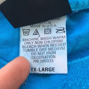 Może przedstawiać: Biała metka pielęgnacyjna z czarnym tekstem i symbolami. Metka zawiera napis "Prać w pralce w ciepłej wodzie, używać tylko wybielacza bez chloru w razie potrzeby, suszyć w suszarce bębnowej w średniej temperaturze, nie prasować dekoracji, RN 105033, XX-Large".