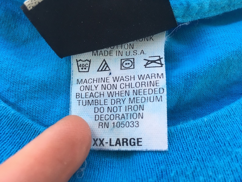 Może przedstawiać: Biała metka pielęgnacyjna z czarnym tekstem i symbolami. Metka zawiera napis "Prać w pralce w ciepłej wodzie, używać tylko wybielacza bez chloru w razie potrzeby, suszyć w suszarce bębnowej w średniej temperaturze, nie prasować dekoracji, RN 105033, XX-Large".