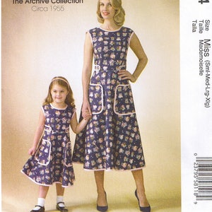 May include: McCall's sewing pattern M7354 featuring a mother and daughter wearing matching dresses. The dresses are navy blue with a floral print and pink trim. The pattern is from The Archive Collection, Circa 1955.