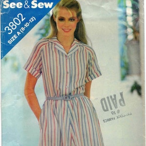 Puede incluir: Sobre de patrón de costura vintage para un vestido a rayas. El vestido presenta rayas verticales en rojo, blanco y azul. El sobre dice "See & Sew" y "Talla A (8-10-12)". El vestido tiene cuello y cintura con cinturón.