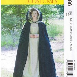 May include: A McCall's Costumes sewing pattern for a hooded cape. The pattern is for sizes X-Small to X-Large. The model is wearing a green velvet cape with a white lace trim and a white dress.