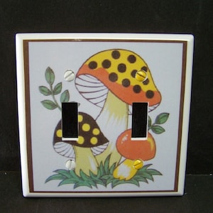May include: A white ceramic light switch cover with a brown border. The cover features a colorful illustration of three mushrooms with red, yellow, and black accents. The mushrooms are surrounded by green leaves.