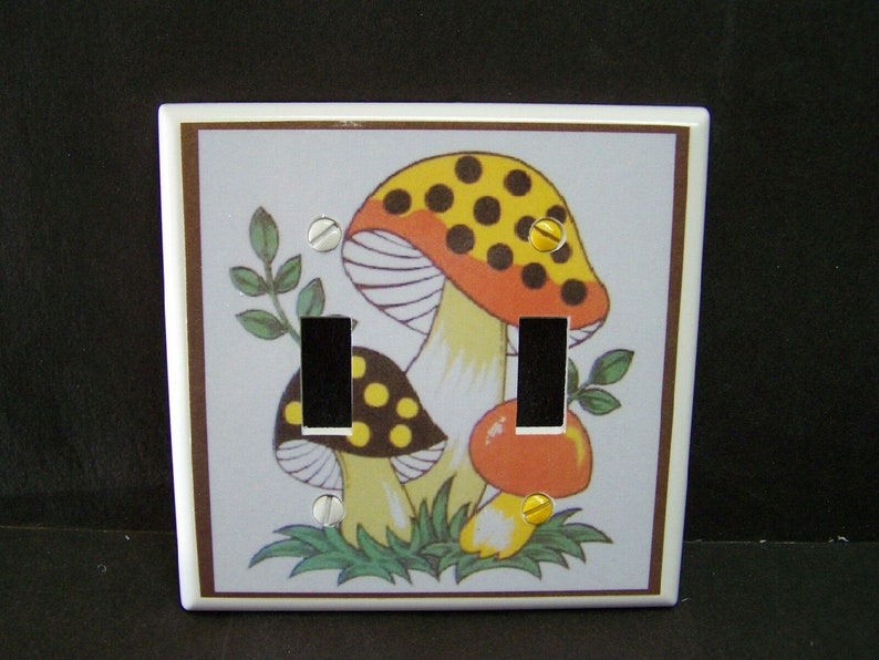 May include: A white ceramic light switch cover with a brown border. The cover features a colorful illustration of three mushrooms with red, yellow, and black accents. The mushrooms are surrounded by green leaves.