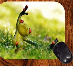 May include: A green frog figurine with red accents, posed in a yoga-like position, sits in a patch of green grass. The frog is on a mousepad with a wooden border, and a black computer mouse is in the foreground.