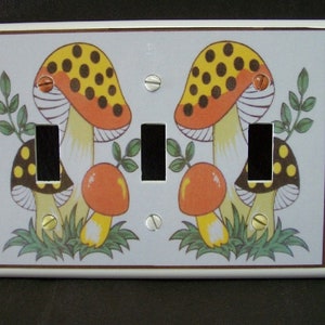 May include: A white ceramic light switch cover with a brown border. The cover features a colorful illustration of two mushrooms with red and orange caps, yellow stems, and black and yellow polka dots. The mushrooms are surrounded by green leaves and grass.