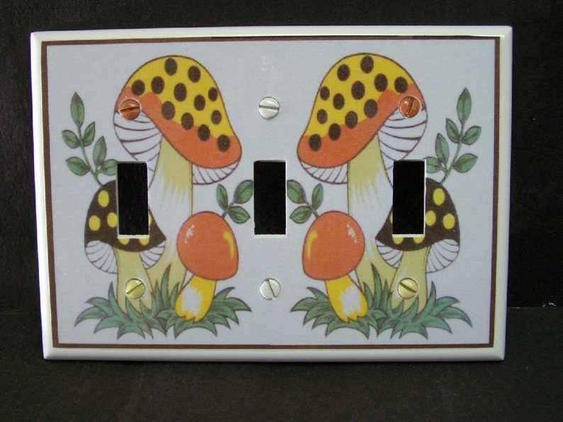 May include: A white ceramic light switch cover with a brown border. The cover features a colorful illustration of two mushrooms with red and orange caps, yellow stems, and black and yellow polka dots. The mushrooms are surrounded by green leaves and grass.