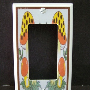 May include: White ceramic light switch cover with a brown border and a design of two yellow and orange mushrooms with brown spots. The mushrooms are surrounded by green leaves.