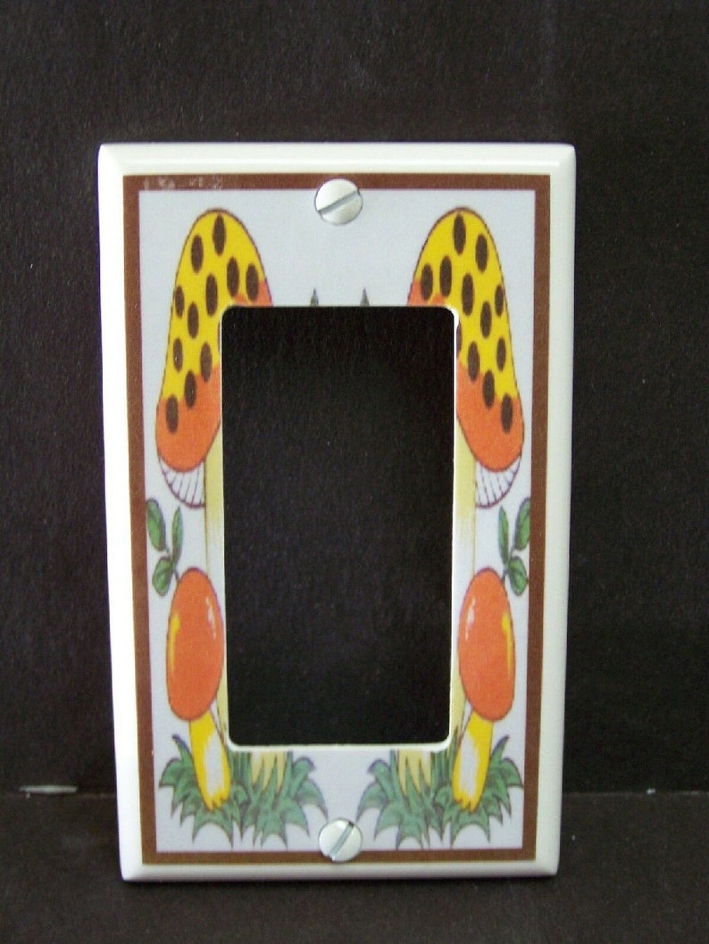 May include: White ceramic light switch cover with a brown border and a design of two yellow and orange mushrooms with brown spots. The mushrooms are surrounded by green leaves.