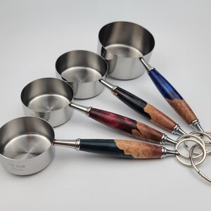 May include: Set of four stainless steel measuring cups with decorative wooden handles. The handles feature a variety of colors, including blue, black, red, and green, with a natural wood grain. The cups are attached with a metal ring.