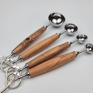 May include: Set of four stainless steel measuring spoons with olive wood handles. The spoons are attached to a metal ring. The handles have a natural wood grain pattern. The spoons are different sizes.