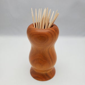 May include: A wooden toothpick holder with a rounded, hourglass shape. The holder is made of a rich, reddish-brown wood and is filled with toothpicks.