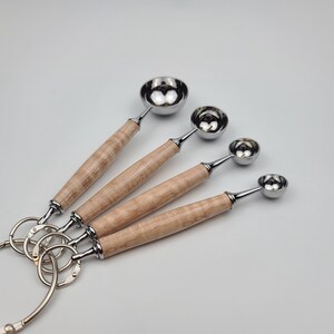 May include: Set of four stainless steel measuring spoons with light wood handles. The spoons are attached to a metal ring. The handles have a light brown and tan wood grain pattern. The spoons are different sizes.