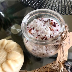 May include: A glass jar containing white bath salts with red petals. A flower-shaped soap and a sage bundle are also present. The jar has a small tag that says "Eucalyptus".