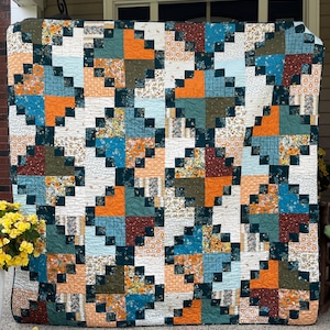 Puede incluir: Un edredón hecho a mano con un diseño de patchwork. El edredón presenta cuadrados en tonos de naranja, verde azulado, verde oscuro y estampados florales, sobre un fondo blanco. El edredón se exhibe al aire libre, mostrando sus colores y texturas.