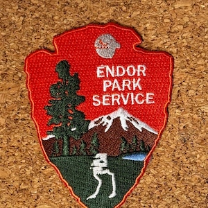 Pode incluir: Patch bordado em forma de ponta de flecha, com o texto "ENDOR PARK SERVICE". O patch tem um fundo vermelho com uma cena de montanha, uma árvore e uma figura branca. O patch é provavelmente para um fã de Star Wars.
