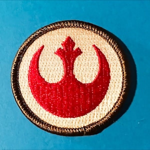May include: A brown and white embroidered patch with the Rebel Alliance symbol in red. The symbol is a crescent moon with a stylized star in the center.