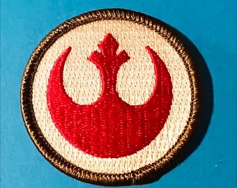 May include: A brown and white embroidered patch with the Rebel Alliance symbol in red. The symbol is a crescent moon with a stylized star in the center.