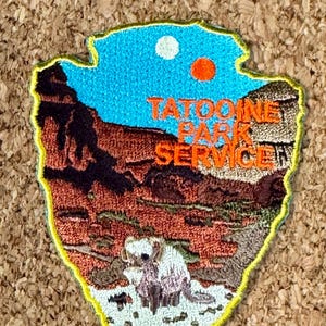 以下が含まれることがあります： 矢印型の刺繍パッチ。砂漠の風景が描かれ、「Tatooine Park Service」の文字が入っています。パッチには、2つの太陽がある青い空と、砂漠の地形の詳細な描写が含まれています。