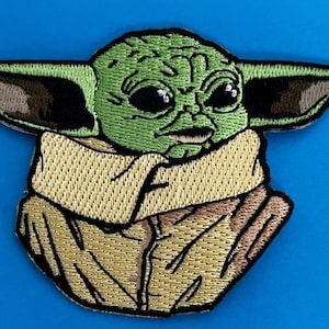 May include: Green and brown embroidered patch of a character from the Star Wars franchise, known as "The Child" or "Baby Yoda". The character is wearing a brown robe and has large ears.
