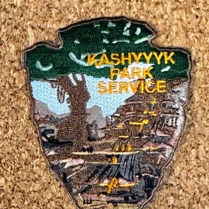 May include: Embroidered patch in the shape of an arrowhead, featuring a landscape scene and the text "Kashyyyk Park Service" in yellow. The patch has a green border and depicts a detailed landscape with trees, mountains, and water.