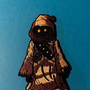 May include: Embroidered patch of a brown robed figure with yellow eyes. The figure is standing with its arms crossed.