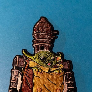 May include: Embroidered patch featuring a green baby Yoda character in a brown pouch, being carried by a gray robot with a red light on top.