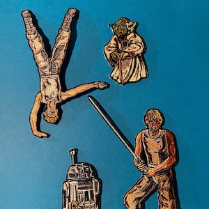 Op de afbeelding: Een set van vier geborduurde patches met personages uit Star Wars. De patches zijn op een blauwe achtergrond. De patches bevatten Luke Skywalker, Yoda, R2-D2 en Luke Skywalker met een lightsaber.
