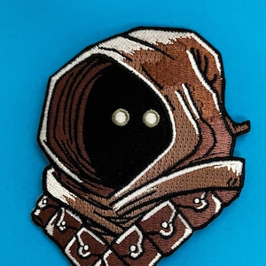 May include: Embroidered patch of a brown hooded figure with white accents and two large white eyes. The figure is wearing a brown and white patterned garment with a brown collar.