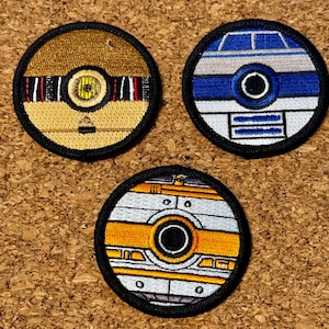 Pode incluir: Três patches redondos bordados com designs de robôs. Um é dourado com detalhes em preto e vermelho, outro é azul e branco, e o terceiro é laranja e prateado. Cada patch tem uma borda preta.